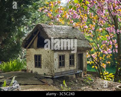 Gros plan d'une maison miniature rustique avec un toit de chaume, entourée de fleurs printanières et de végétation verte dans un cadre de jardin. Banque D'Images
