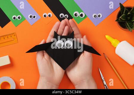 Mains féminines tenant la batte jouet en papier origami fini. Sur la table se trouvent des outils pour papier déplaçables Concept de projet DIY pour les enfants. Pose à plat Banque D'Images