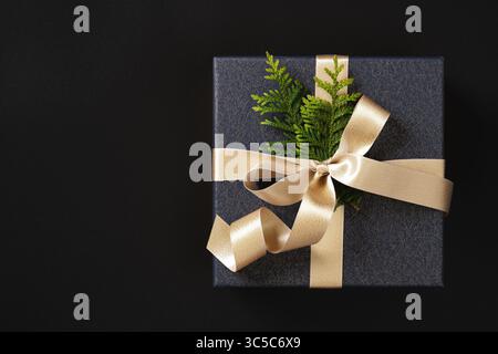 Le coffret cadeau en papier bleu foncé élégant est attaché avec un ruban de satin doré et décoré avec une branche de thuja. Fond noir, espace pour votre Banque D'Images