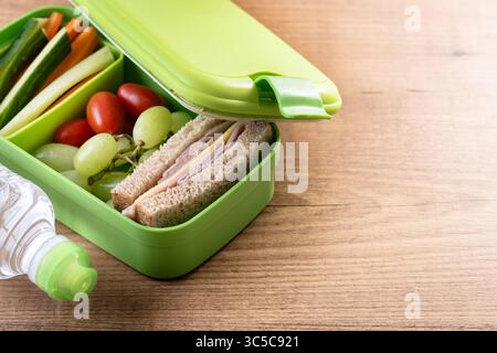 Gros plan d'une boîte à lunch verte sur une table en bois, contenant un sandwich au jambon et au fromage, des raisins, des tomates cerises et des bâtonnets de légumes Banque D'Images