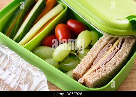 Gros plan d'une boîte à lunch verte sur une table en bois, contenant un sandwich au jambon et au fromage, des raisins, des tomates cerises et des bâtonnets de légumes Banque D'Images
