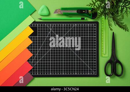 Un tapis de découpe noir avec des lignes de grille blanches sur un fond vert vif, entouré d'articles de papeterie soigneusement disposés : un crayon, des ciseaux, un cutter, Banque D'Images