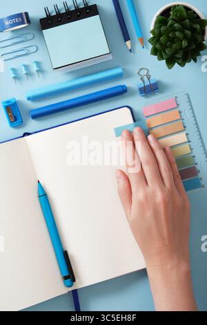 Vue de dessus d'un bureau soigné. Une main lisse un autocollant collé dans un carnet ouvert avec une page blanche. Autour des stylos bleus de fournitures de bureau, des crayons, du pe feutre Banque D'Images