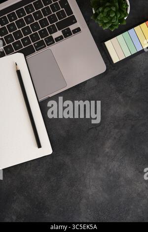 Espace de travail minimaliste avec ordinateur portable, planificateur de balles, stylo, autocollants et plante succulente sur un fond texturé sombre. Copier l'espace. Banque D'Images