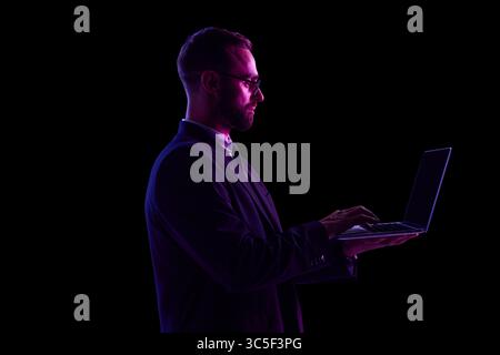 Homme en costume sombre utilisant un ordinateur portable dans l'éclairage au néon sur fond noir Banque D'Images