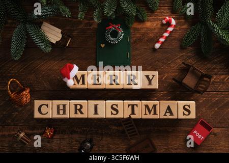 Blocs en bois avec texte joyeux Noël en anglais avec des branches d'arbre de Noël et des détails mignons, chaise à bascule, boîte aux lettres, porte avec couronne, chapeau de Père Noël Banque D'Images