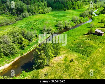 Une perspective aérienne capture une rivière sereine serpentant à travers des champs verdoyants, avec des arbres dispersés et des structures rurales lointaines Banque D'Images