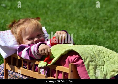 Jeune fille allongée dans un lit de jouet sur l'herbe, enveloppée dans une couette verte et portant des vêtements rayés. Jouet joyeux en plein air sous la lumière du soleil. Banque D'Images