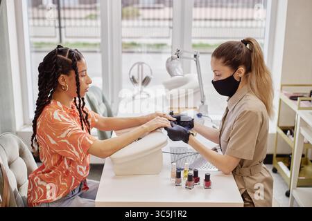 Biracial jeune femme adulte recevant la manucure de femme caucasienne d'âge moyen portant un masque de protection et des gants, tous deux assis à la table de salon de ongles wi Banque D'Images