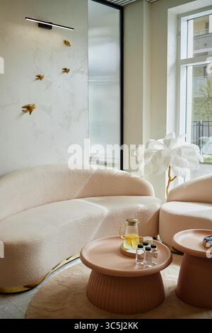 Salle d'attente moderne dans le salon de beauté avec canapé beige courbé, tables basses rondes avec pichet en verre d'eau et petites bouteilles, mur décoratif Ar Banque D'Images