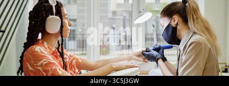 Capture d'en-tête du site Web d'une jeune femme biraciale adulte portant des écouteurs recevant une manucure d'une jeune femme caucasienne adulte avec queue de cheval et masque facial i. Banque D'Images