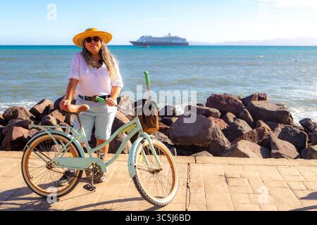 Touriste féminine debout à côté de vélo sur la promenade, la mer et le bateau de croisière en arrière-plan, portant un short, chemisier blanc, chapeau et lunettes de soleil, jour ensoleillé i Banque D'Images