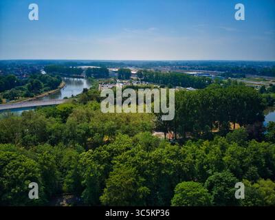 Vue aérienne de la canopée verte de la forêt rencontre des voies navigables sinueuses sous un vaste ciel bleu, encadré par des bâtiments lointains, Bruges, Flandre, Belgique. Banque D'Images