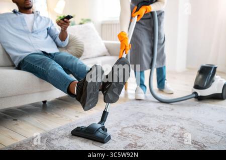 Femme noire passant l'aspirateur sur le sol pendant que son mari regarde la télévision, ne l'aidant pas avec les tâches ménagères, vue rognée Banque D'Images