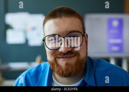 Portrait de jeune homme caucasien adulte avec les cheveux courts et la barbe souriant et regardant dans la caméra, portant des lunettes, assis à l'intérieur avec un fond de bureau flou Banque D'Images