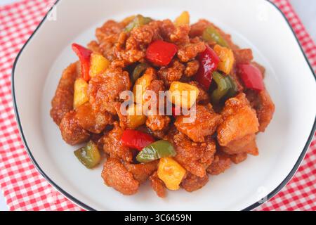 Gros plan de poulet aigre-doux fait maison avec poivrons et morceaux d'ananas, servi sur une assiette blanche avec une serviette à carreaux rouge. Banque D'Images