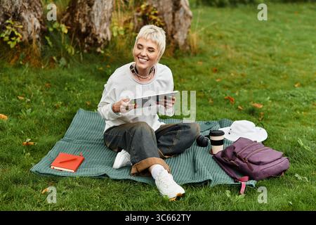 Charmante jeune femme se détend sur une couverture dans le parc, souriant et utilisant sa tablette avec joie. Banque D'Images