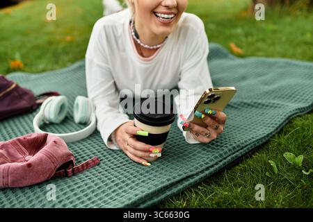 Une jeune femme élégante se détend dans un parc, sirotant un café tout en utilisant son téléphone, souriant joyeusement. Banque D'Images