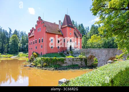 Le château Cervena Lhota est situé sur une île rocheuse au centre d'un lac, avec des murs rouges vibrants et une architecture historique au milieu d'un paysage verdoyant en Tchéquie. Banque D'Images