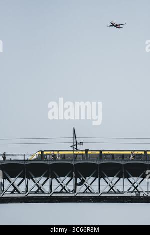 Porto, Portugal - 17 juin 2022 : vue d'un train jaune moderne traversant le pont métallique Dom Luís I alors qu'un avion vole au-dessus, un mélange de modes de transport dans un ciel silencieux. Banque D'Images