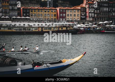 Porto, Portugal - 17 juin 2022 : vue sur le fleuve Douro reflétant le quartier coloré de Ribeira, avec des rameurs glissant devant des bateaux traditionnels sous un ciel couvert. Banque D'Images