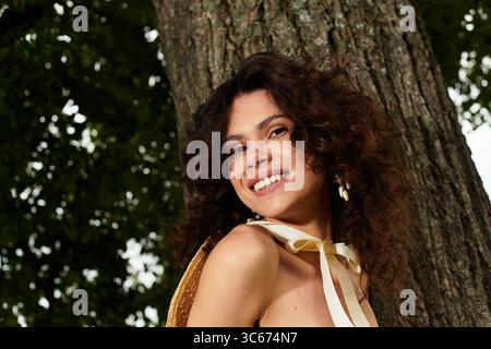 Belle jeune femme sourit joyeusement tout en étant entourée de verdure luxuriante dans un parc de village. Banque D'Images