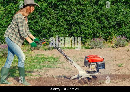 Fermière en chapeau et jeans laboure le sol avec cultivateur à essence à main dans le jardin, préparant la terre pour l'ensemencement. Concept agricole. Banque D'Images