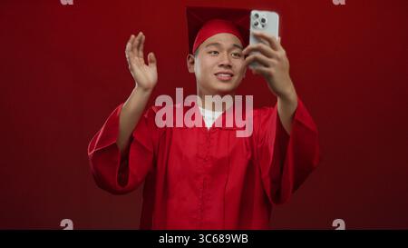 Jeune homme chinois étudiant en robe de graduation rouge faisant un appel vidéo sur smartphone au-dessus d'un mur rouge isolé célébrant la réussite scolaire avec une joyf Banque D'Images