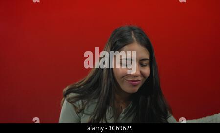 Jeune femme joue de la guitare à air avec les mains contre le mur rouge du studio tout en souriant et en se penchant en avant ; joie ludique. Banque D'Images