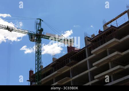 Vue en angle bas d'un bâtiment en béton en construction avec une grue à tour verte contre un ciel bleu vif avec des nuages. Banque D'Images