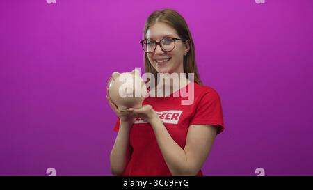 Jeune femme blonde portant une chemise de bénévole souriant tient une tirelire en studio ; générosité don de charité. Banque D'Images