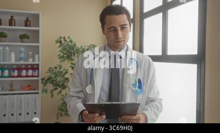 Jeune homme médecin en manteau blanc tient une presse-papiers et lit le dossier du patient dans la salle de la clinique par fenêtre ; dévouement. Banque D'Images