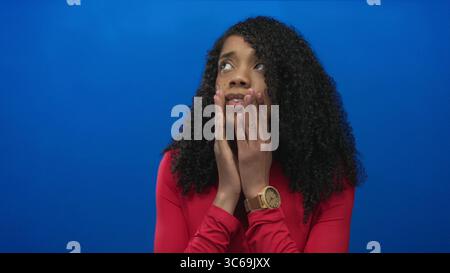 Femme afro-américaine avec les mains serrées près des lèvres dans le studio bleu, les yeux fermés dans l'expression inquiète ; anxiété. Banque D'Images