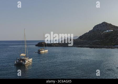 Vue de deux voiliers blancs reposant sur les eaux immobiles et bleu foncé près d'une côte rocheuse sous un ciel clair et bleu pâle, Kallithea, Rodhes, Grèce. Banque D'Images