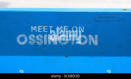 Le texte au pochoir blanc « MEET ME ON OSSINGTON » est peint sur une barrière de béton bleu vif le long de la rue. Banque D'Images