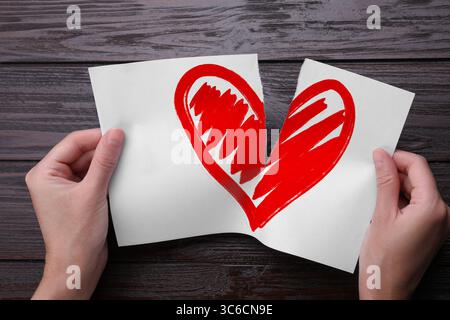 Trahison. Femme déchirant la feuille de papier avec le coeur rouge en moitiés sur fond de bois, gros plan Banque D'Images