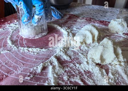 Les mains couvertes de gants bleus appuient sur un verre tandis que la poussière de farine tourbillonne autour. La table contient de la pâte fraîchement préparée, révélant la joie de la cuisson Banque D'Images