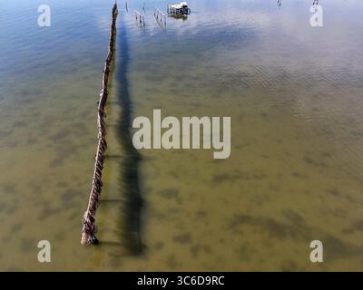 Vue aérienne de poteaux en bois altérés debout dans les eaux peu profondes et réfléchissantes, créant des ombres dures sur le fond sablonneux, Kalochori, Grèce. Banque D'Images