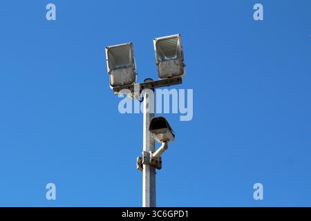 Deux grands projecteurs extérieurs et une caméra de sécurité sont montés sur un haut poteau métallique, assurant la surveillance et l'éclairage contre un bleu vif Banque D'Images