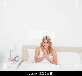 Femme assise et souriante sur le lit à la maison avec lampe de chevet blanche, livre rose, horloge argentée. Détente, confort, sérénité, minimalisme, bien-être, plus vif Banque D'Images