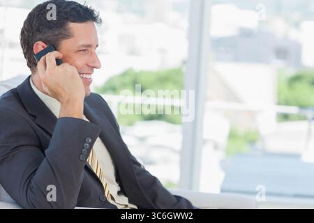 Homme assis dans le salon d'affaires portant costume et cravate tenant smartphone à la fenêtre, espace photocopie. Professionnel, affaires, entreprise, occasionnel, moderne, urba Banque D'Images