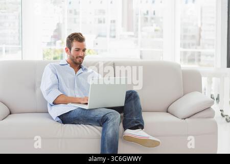 Homme assis sur le canapé gris clair dans le salon lumineux de l'appartement de ville travaillant sur ordinateur portable. Contemporain, professionnel, cosy, minimaliste, productivité, urb Banque D'Images