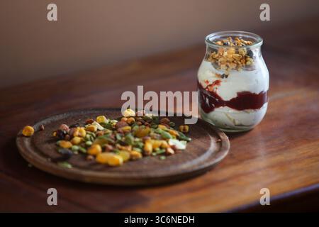 gros plan de pot en verre rempli de yaourt crémeux et d'un mélange en couches de granola croquant et de muesli, placé sur une table en bois avec des noix et des graines dispersées Banque D'Images