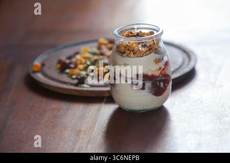 gros plan de pot en verre rempli de yaourt crémeux et d'un mélange en couches de granola croquant et de muesli, placé sur une table en bois avec des noix et des graines dispersées Banque D'Images