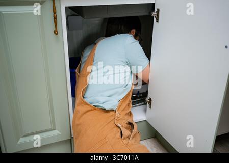 Le plombier professionnel portant une combinaison brune répare les tuyaux du système d'alimentation en eau, connectant le siphon sous l'évier de la cuisine dans un appartement moderne Banque D'Images