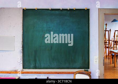 Un vieux tableau vert monté sur un mur de couleur claire à côté et une porte menant à une salle de classe remplie de chevalets et de tabourets. Banque D'Images