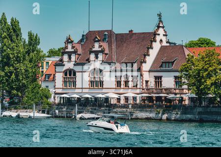Charmant bâtiment ancien avec toit orné et terrasse au bord de l'eau sur les rives du lac de Constance à Constance en Allemagne. Un petit bateau à moteur passe sur un soleil Banque D'Images