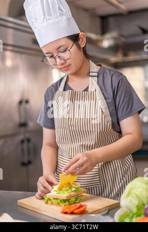 Une jeune femme asiatique, dans la vingtaine, portant un tablier rayé, prépare méticuleusement un sandwich maison délicieux et sain avec des ingrédients frais en HE Banque D'Images