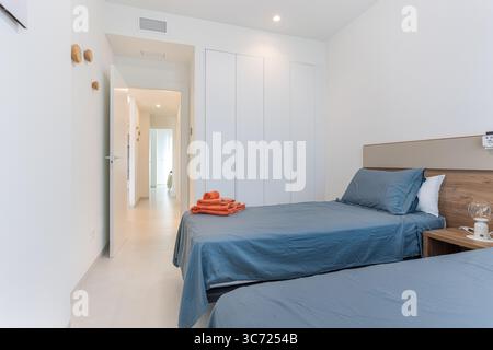 Chambre propre et moderne avec draps bleus, serviettes orange et rangement intégré avec accès au couloir. Banque D'Images