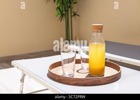 Deux verres et une bouteille de jus d'orange sur un plateau en bois à côté de chaises longues, baignés de soleil et stylisés pour les loisirs de vacances. Banque D'Images